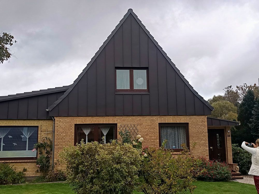 Haus mit braunem Dach und Backsteinverkleidung, einem dreieckigen oberen Teil und einer Person, die in der rechten Ecke ein Foto macht.