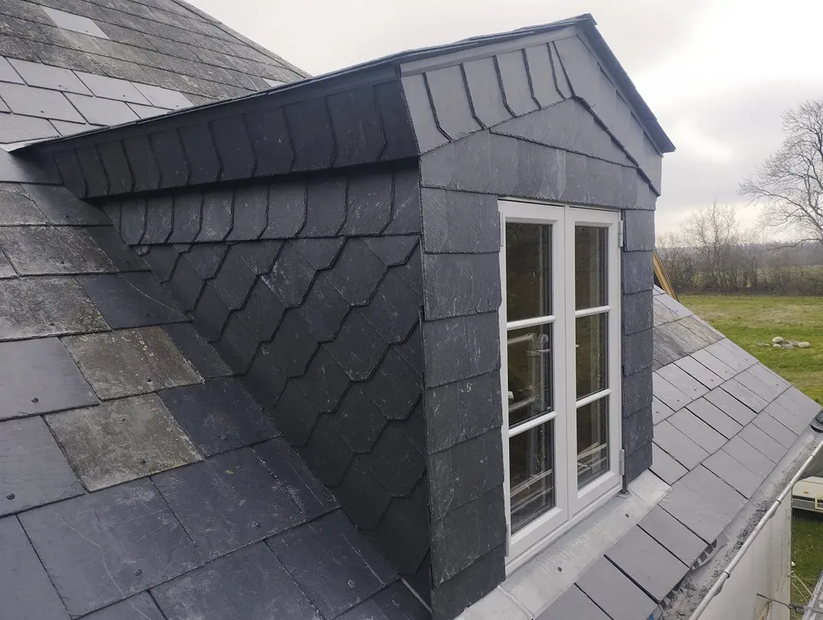 Dachgaubenfenster mit dunkelgrauen Schieferziegeln an einem Haus. Bewölkter Himmel.