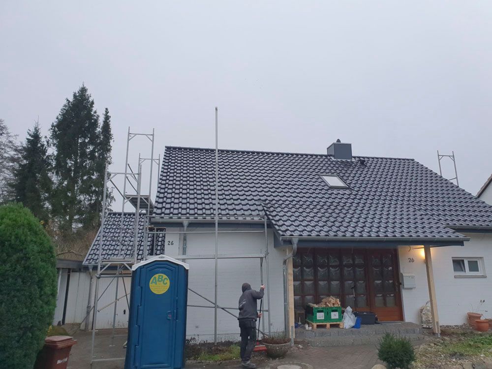 Haus mit neuem, schwarzem Dach, Gerüst und einer mobilen Toilette. Jemand arbeitet an dem Gebäude an einem bewölkten Tag.