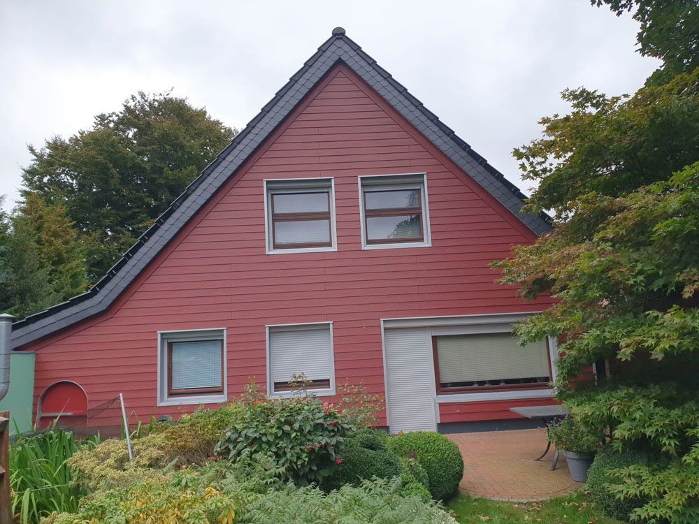 Ein rot verkleidetes Haus mit schwarzem Dach, schwarzen Fenstern und umgebender Vegetation unter bewölktem Himmel.