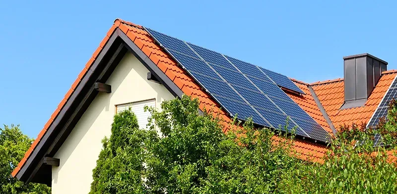 Haus mit Solarpaneelen auf einem roten Ziegeldach, umgeben von Bäumen und einem klaren blauen Himmel.