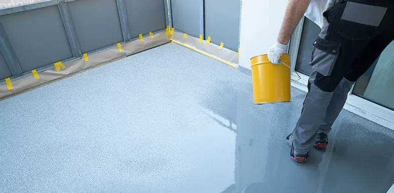 Eine Person gießt graues Epoxidharz aus einem gelben Eimer.