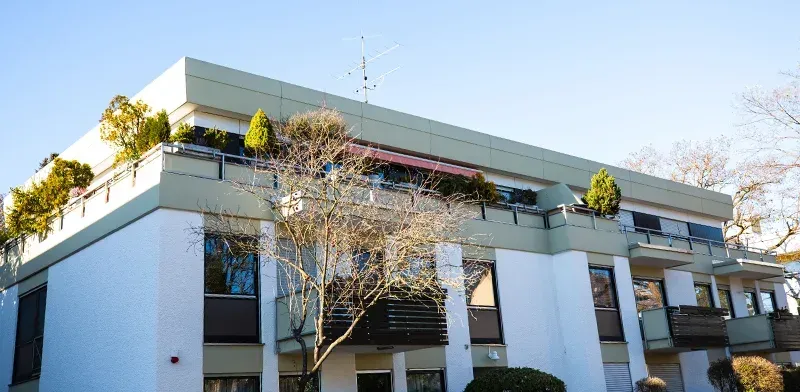 Ein Wohnhaus mit weiß-beigen Wänden, kleinen Bäumen auf dem Balkon und blauem Himmel.