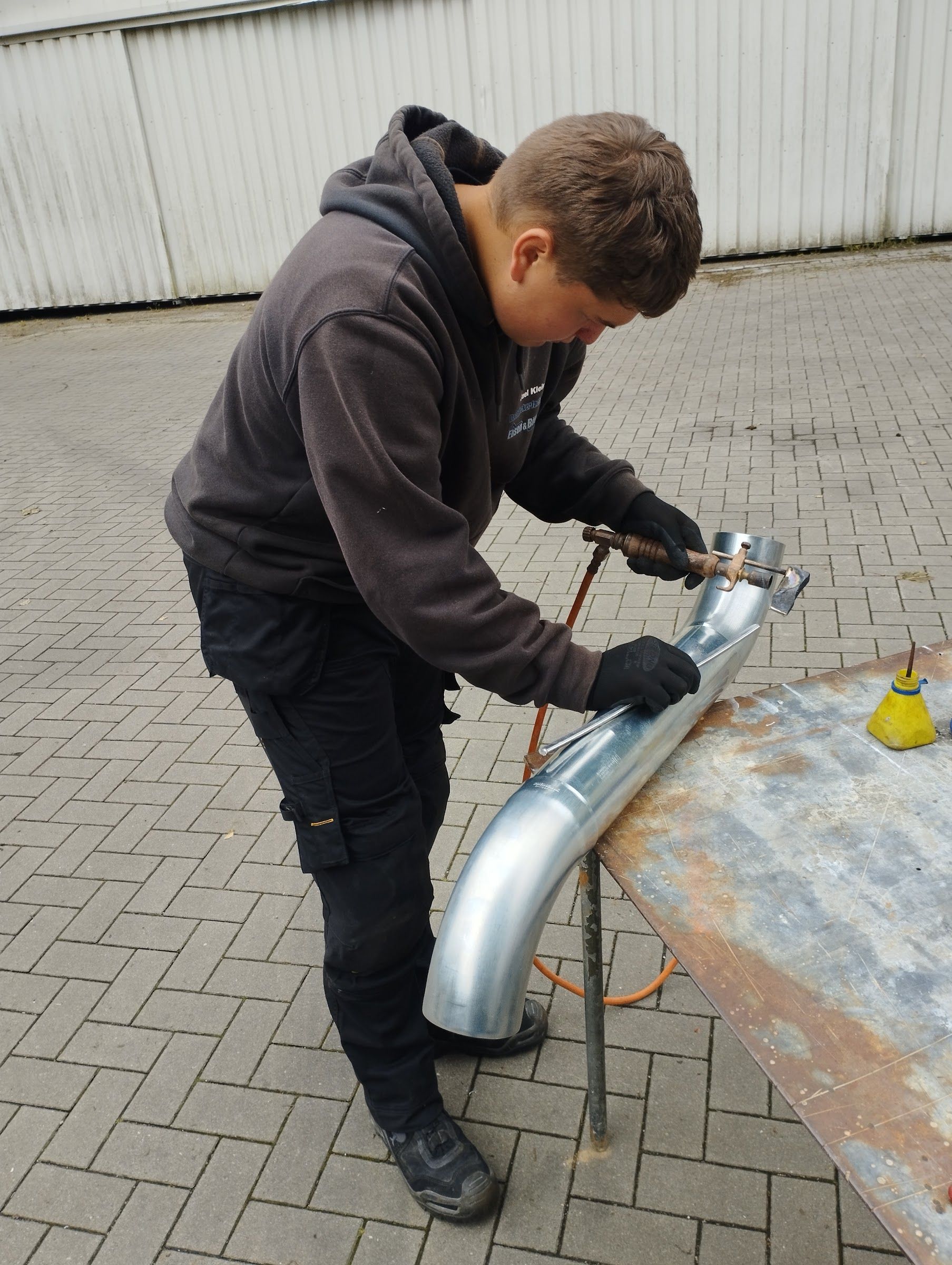 Ein Arbeiter legt einen orangefarbenen Dachziegel auf ein rotes Ziegeldach; im Vordergrund ist eine silberne Dachrinne zu sehen.