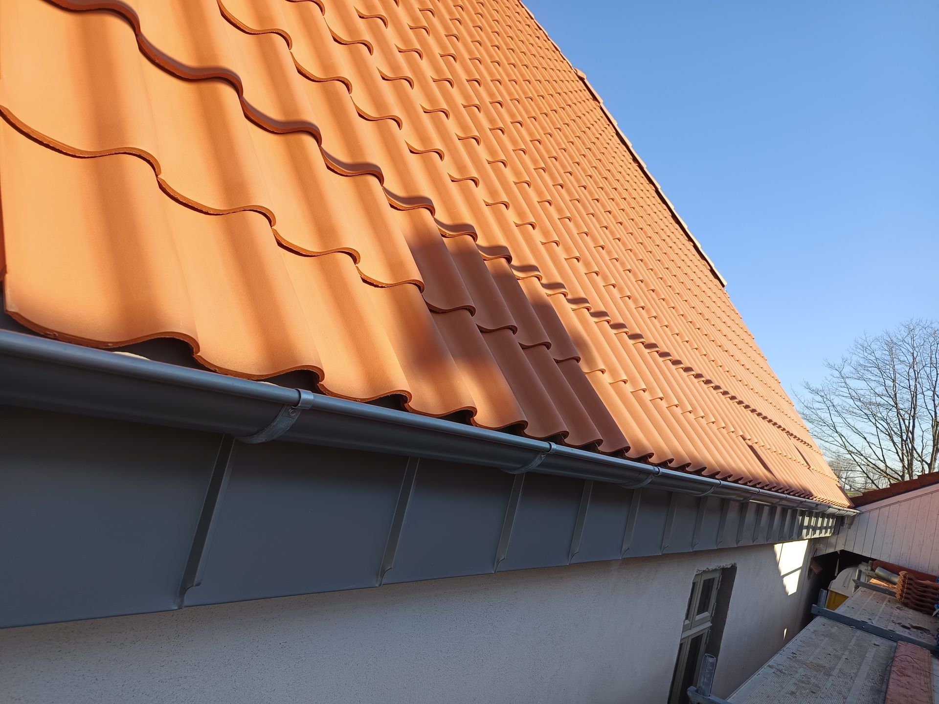 Ein Dachdecker geht auf einem mit Ziegeln gedeckten Dach. Schwarze Hose, Stiefel, im Hintergrund blauer Himmel.