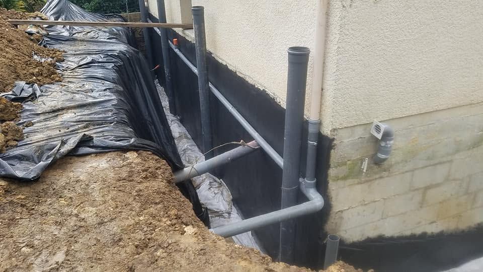 Réparation des fondations avec une membrane d’étanchéité noire et des tuyaux apparents le long d’un mur de maison en stuc.