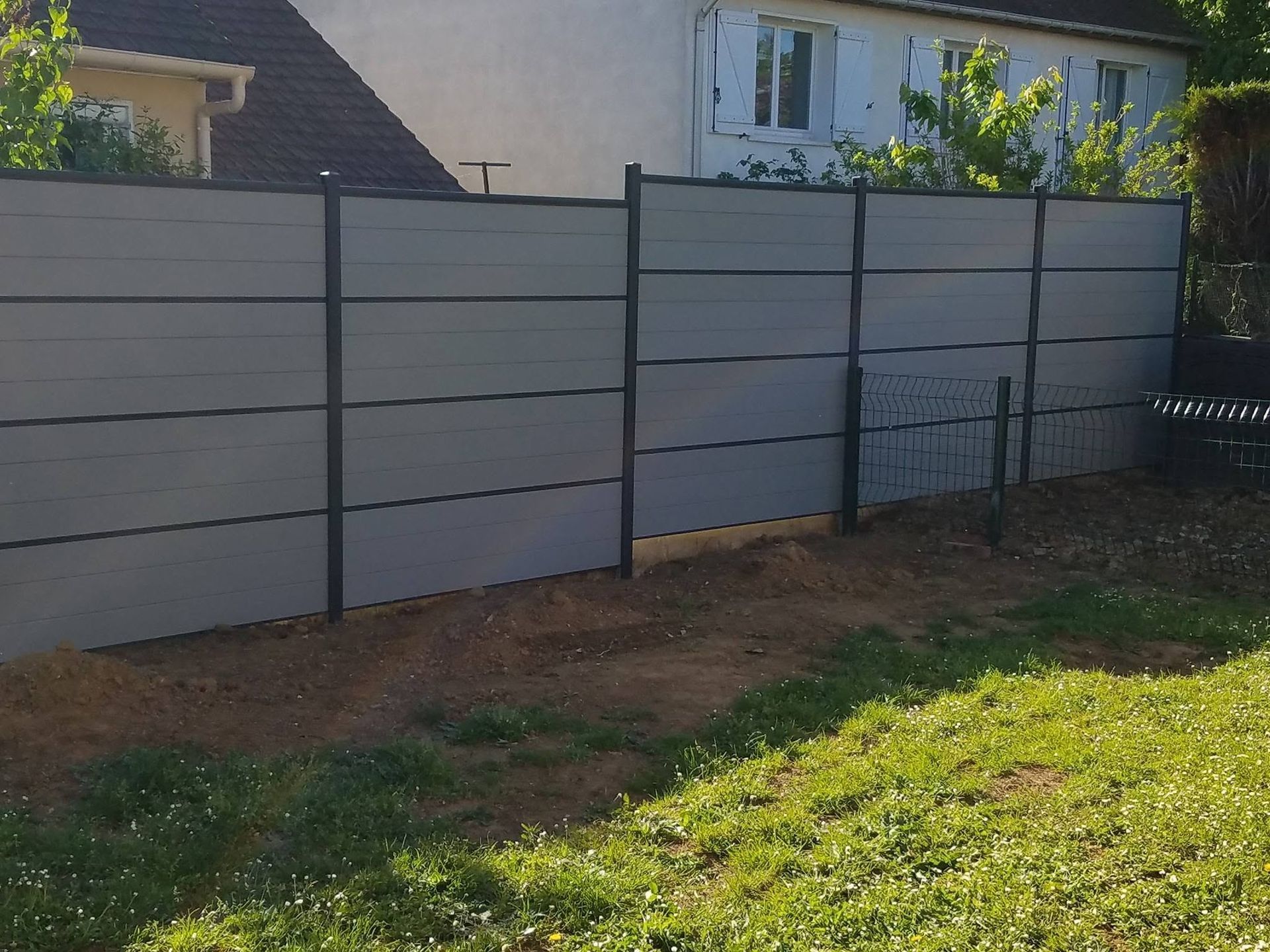 Clôture grise à panneaux dans un jardin, avec un portail, une pelouse verte et une maison en arrière-plan.