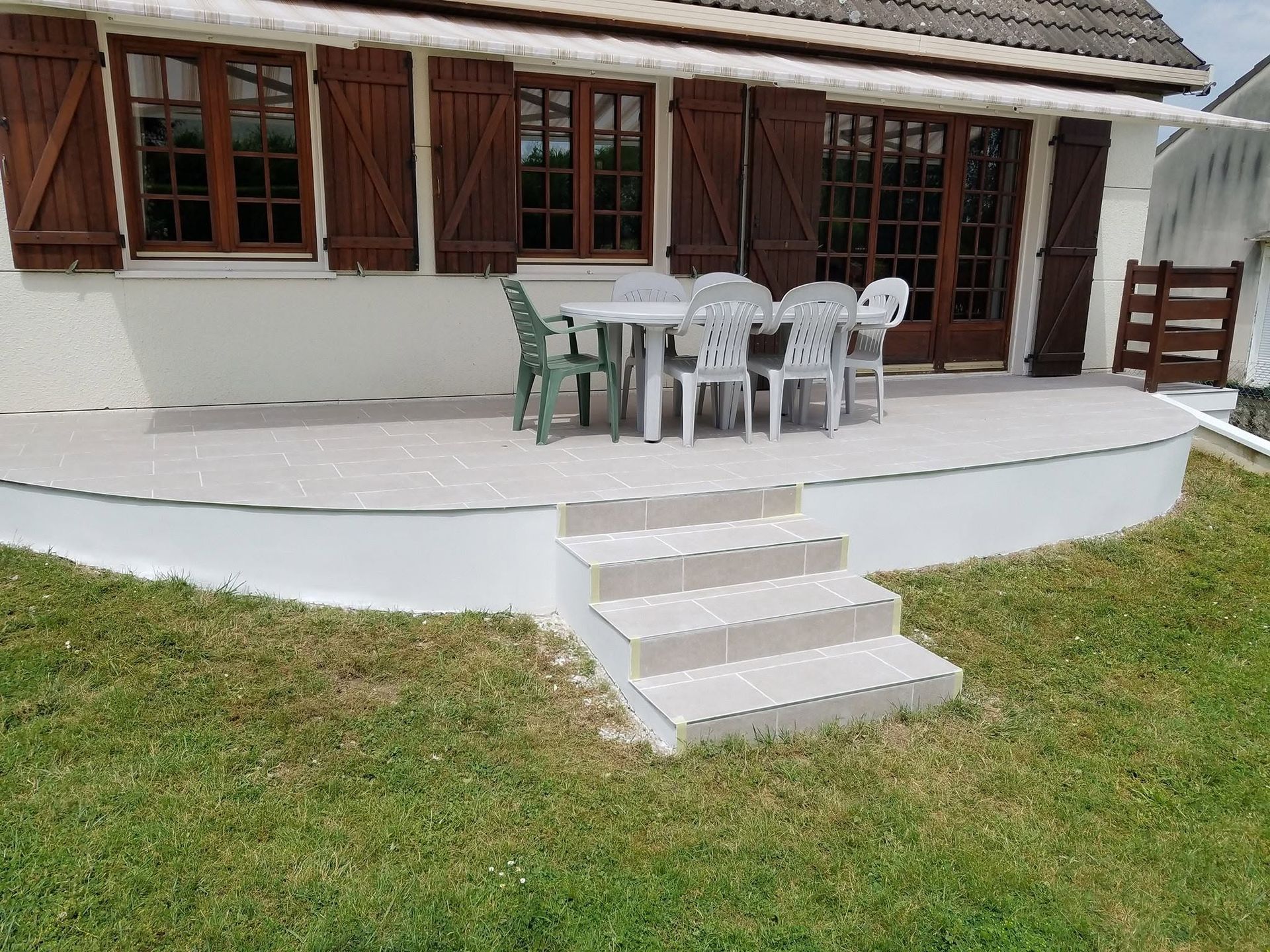 Patio avec une table et des chaises blanches sur une terrasse blanche, quelques marches et une pelouse devant une maison.