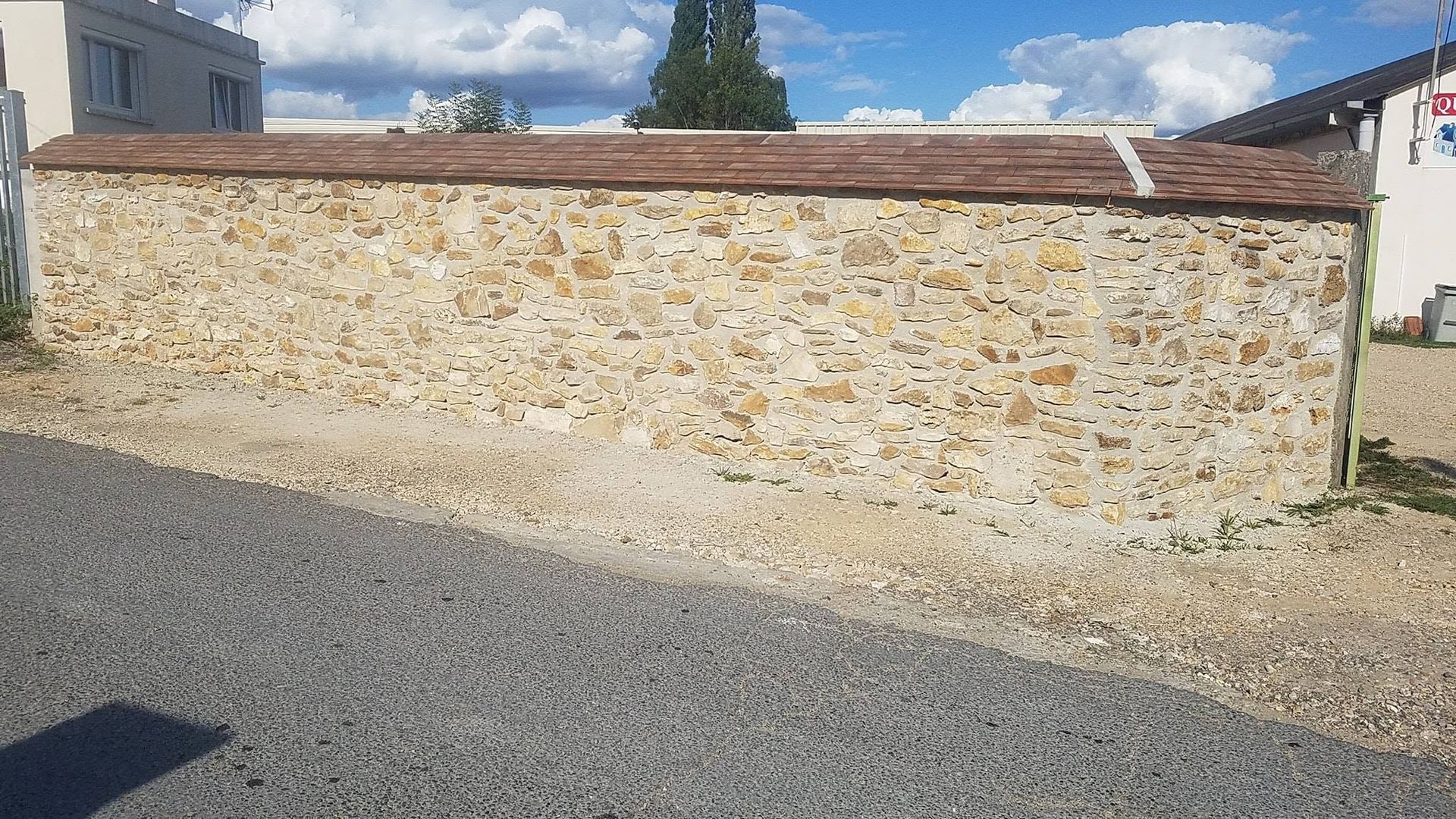 Mur de pierre surmonté de tuiles, au bord d’une route de gravier sous un ciel partiellement nuageux.