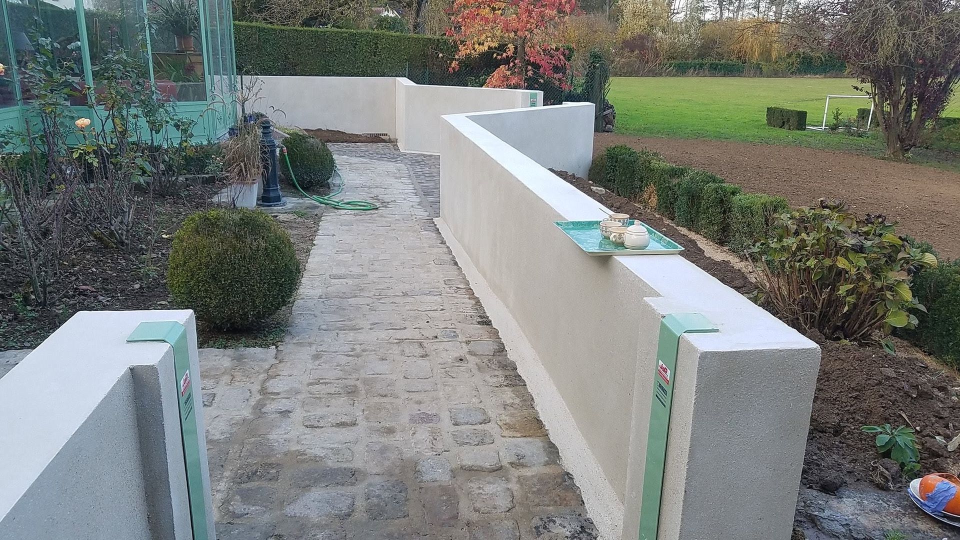 Allée de jardin en pierre entre de petits murs couleur crème, traversant des arbustes et de la verdure en plein air.