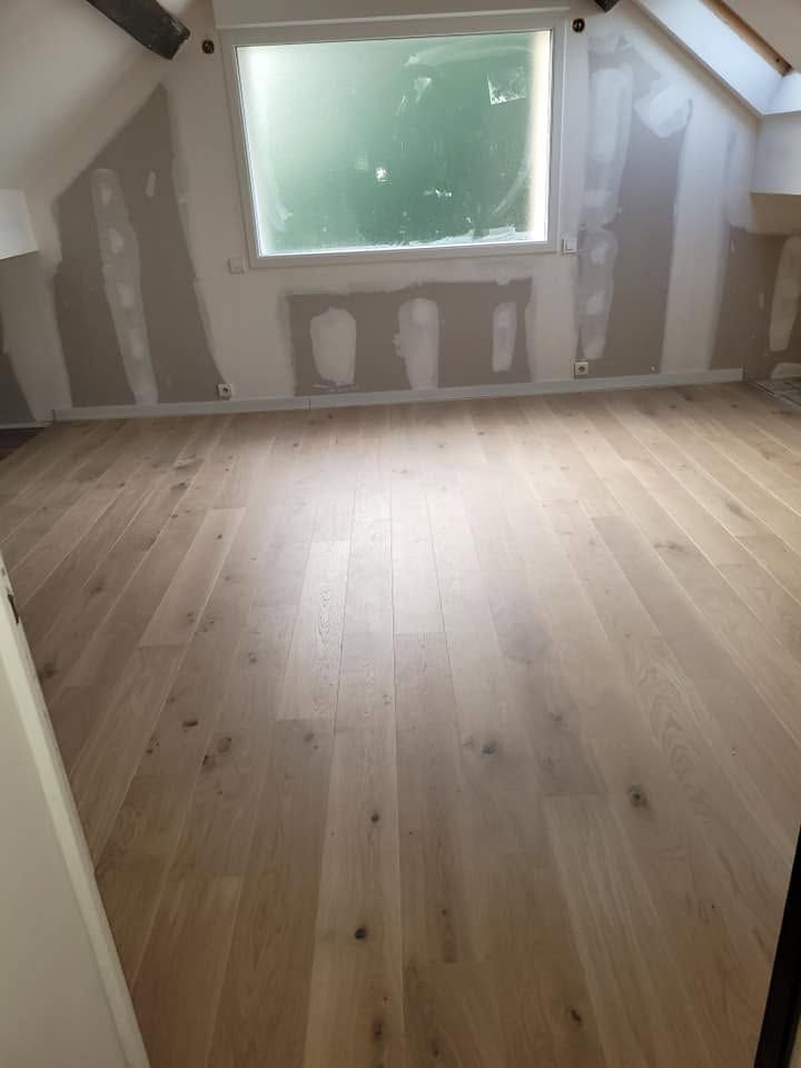 Petite pièce mansardée inachevée, avec des murs blancs en pente, une fenêtre et un parquet clair.