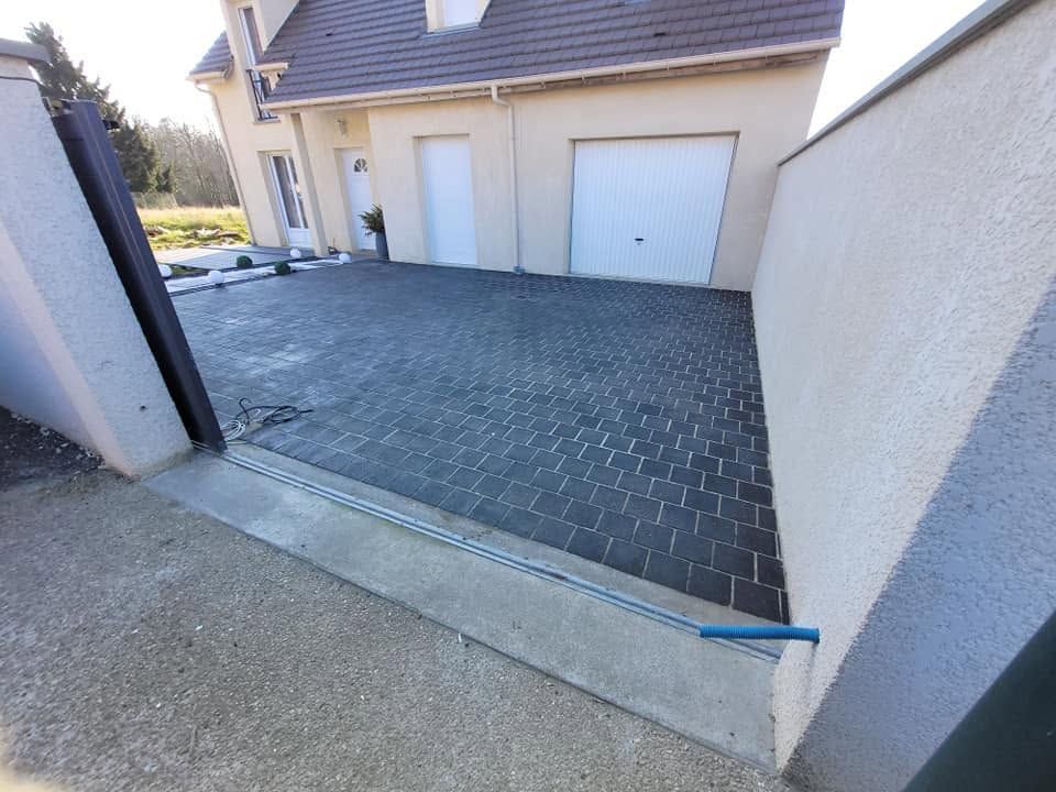 Allée bordée de pavés foncés menant à un garage, à côté d’une maison beige et de murs blancs.