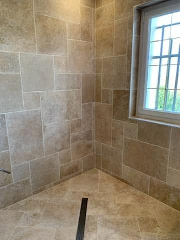 Douche à l’italienne carrelée dans un angle, avec des murs en pierre beige, une fenêtre et une évacuation linéaire au sol.