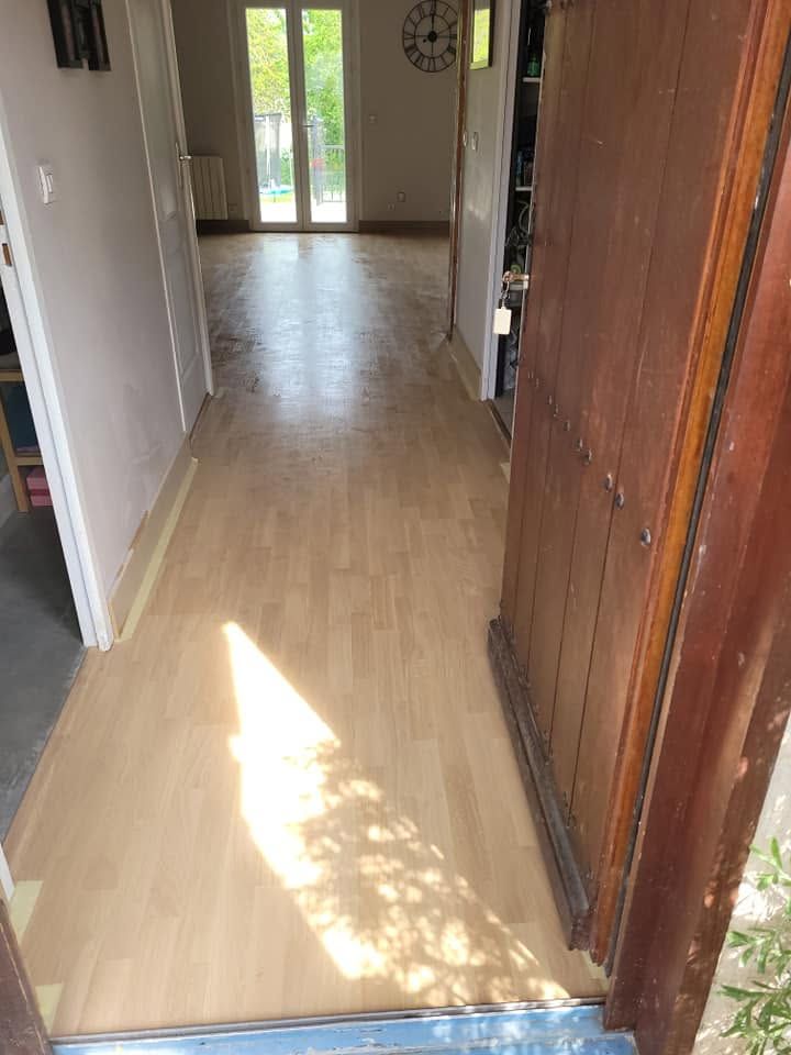 Couloir avec un parquet clair, une zone ensoleillée et une porte d’entrée ouverte donnant sur une porte vitrée au fond.