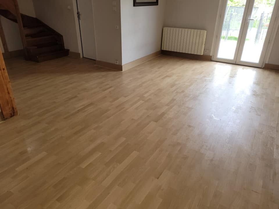Salon vide avec parquet clair, escalier, radiateur et portes vitrées laissant entrer la lumière du soleil.