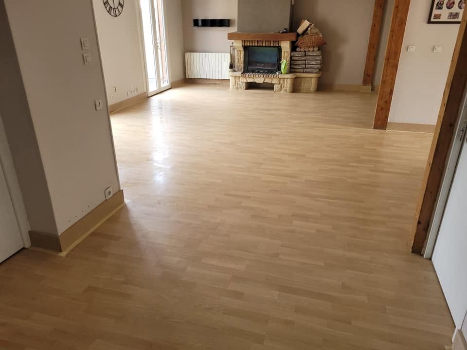 Salon vide avec parquet clair, cheminée et porte sur la droite.