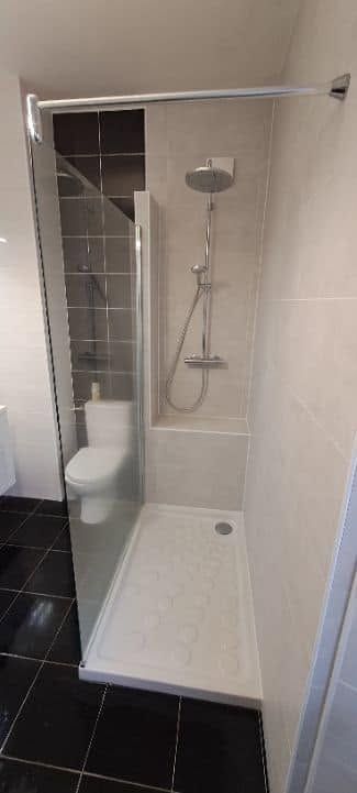 Petite cabine de douche carrelée avec porte vitrée, douchette à main et siège rabattable dans une salle de bains.