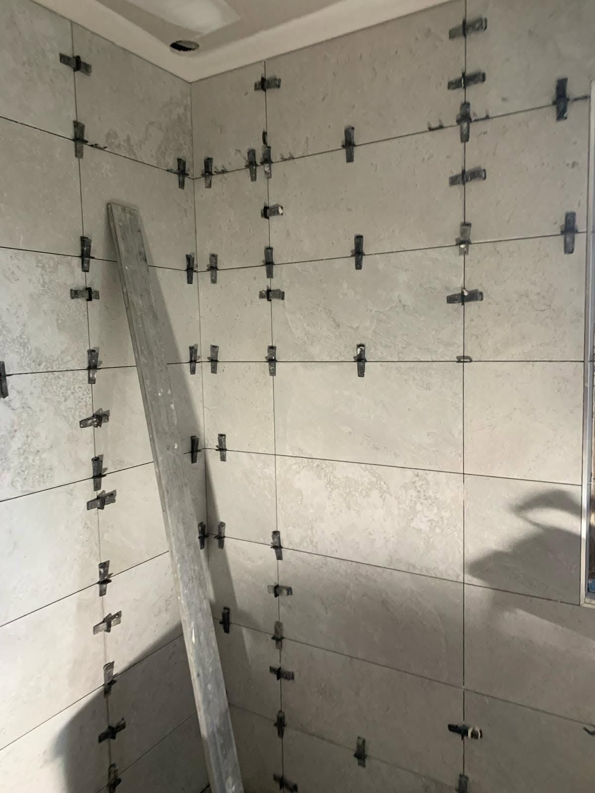 Mur de douche partiellement carrelé, avec des clips de nivellement et un panneau de carrelage incliné pendant la pose.