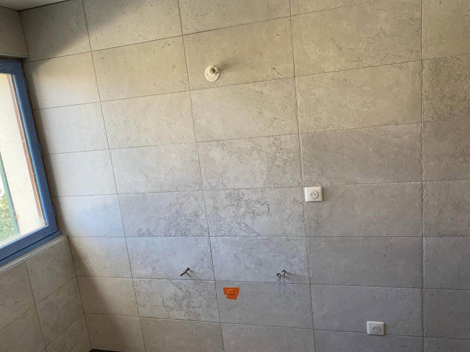 Mur de salle de bains carrelé et vide, avec une fenêtre, des prises électriques et un petit autocollant orange.