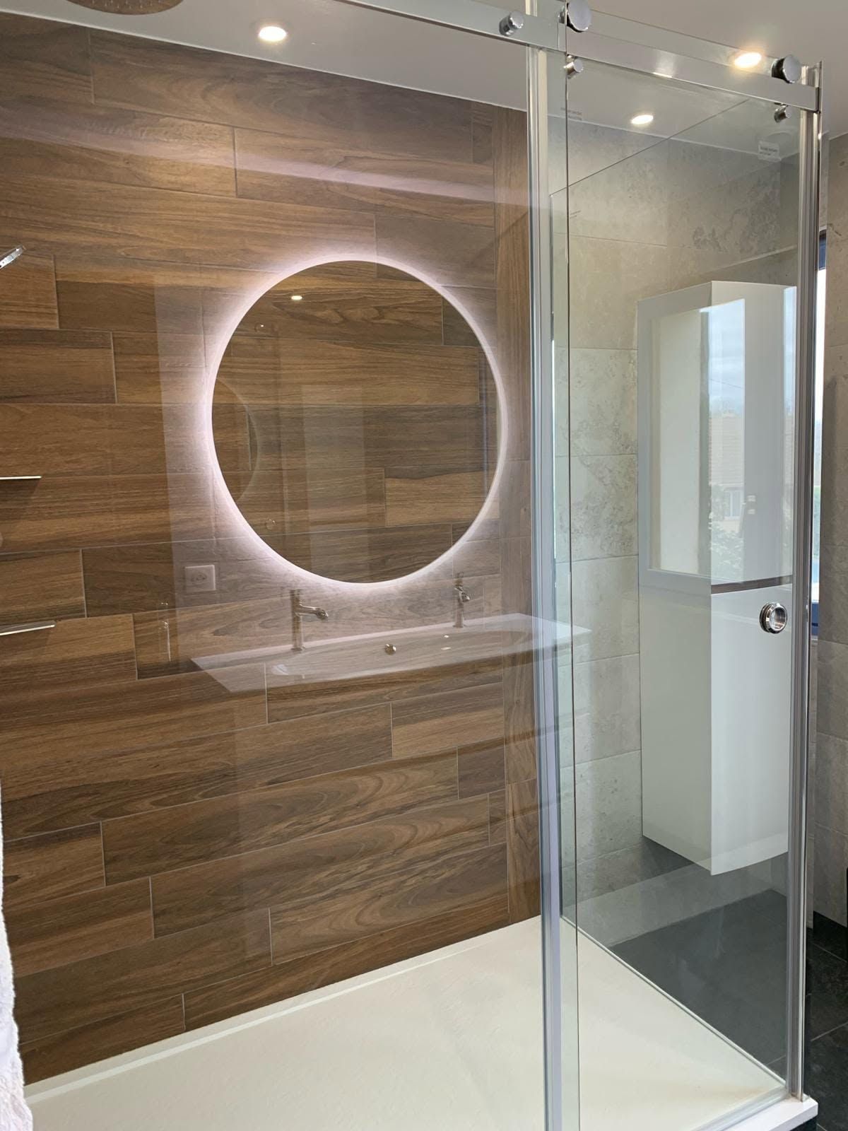 Salle de bains moderne avec douche vitrée, mur lambrissé de bois et miroir rond éclairé.
