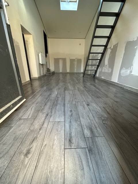 Intérieur de loft vide avec un parquet gris, une échelle menant à la mezzanine et une fenêtre de toit au-dessus.