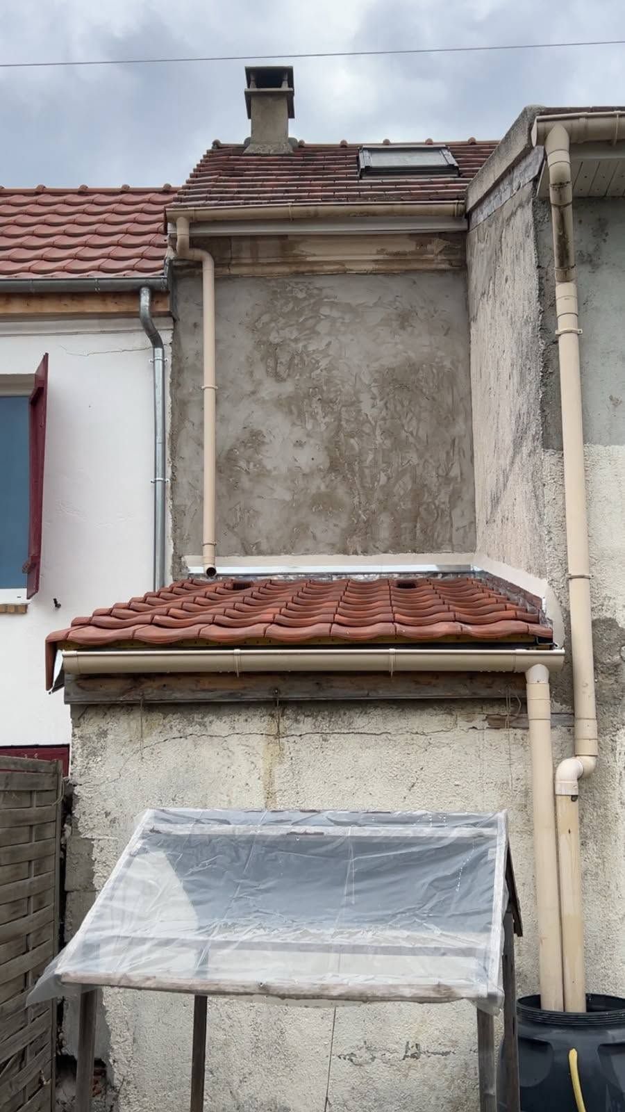 Façade étroite d’une maison crépie, avec un toit de tuiles rouges et un auvent couvert, vue entre les bâtiments voisins.