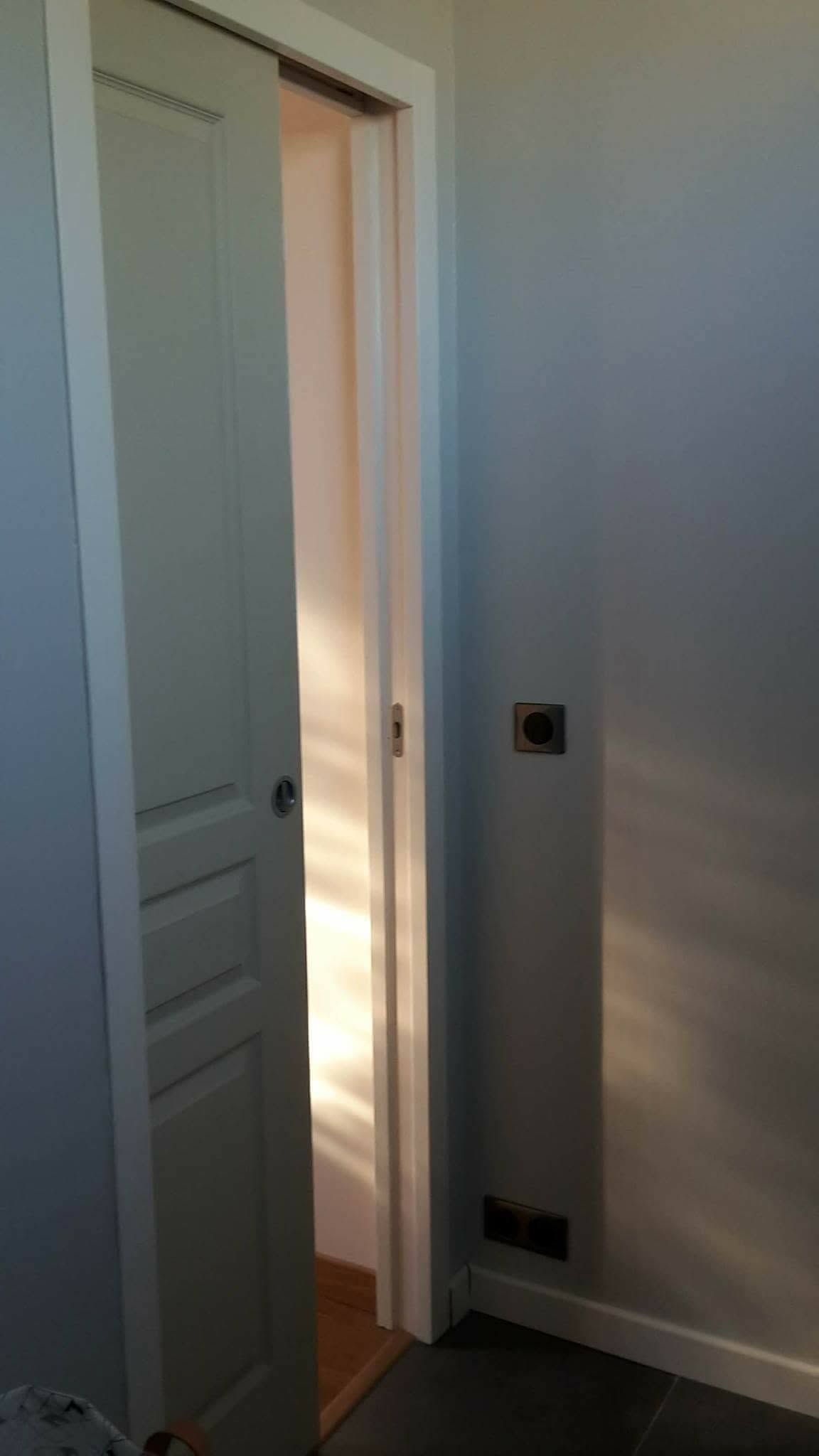 Une porte blanche, laissant une lumière chaleureuse se répandre dans un couloir blanc ; une prise murale est visible à proximité.