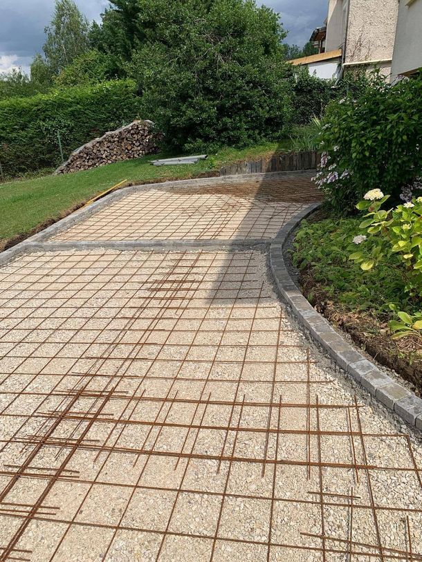 Allée en pierre incurvée en cours de construction, avec un treillis de renforcement, bordée d’herbe et d’arbustes.