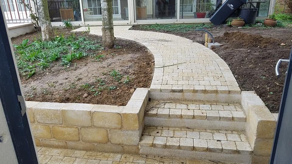 Allée pavée de briques incurvée, avec des marches menant à l’entrée d’un bâtiment, bordée de massifs paysagers et d’arbres.