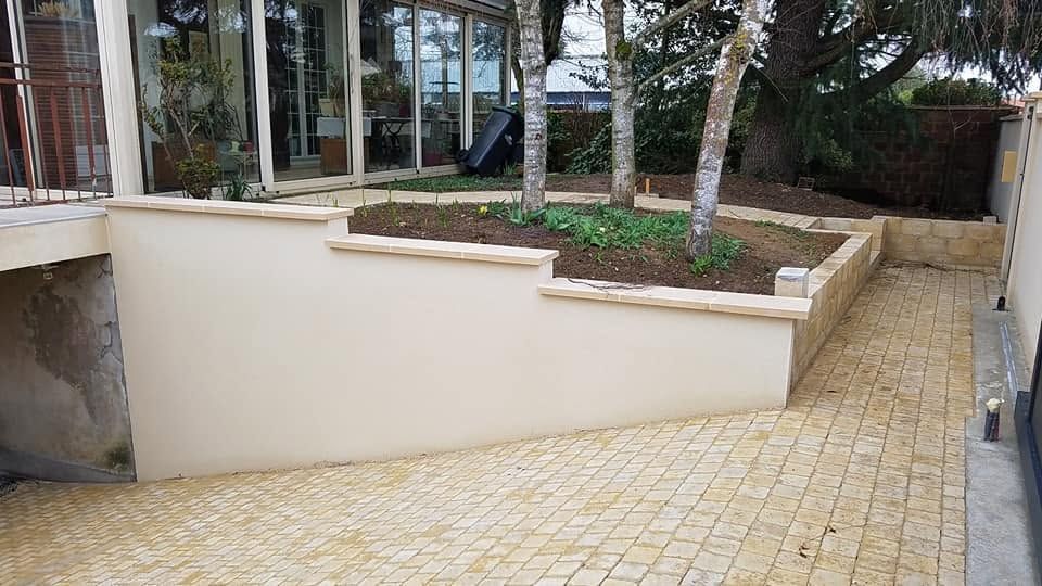 Patio extérieur avec une rampe beige à côté de bacs de plantation surélevés et d’arbres, près de l’entrée d’un bâtiment.