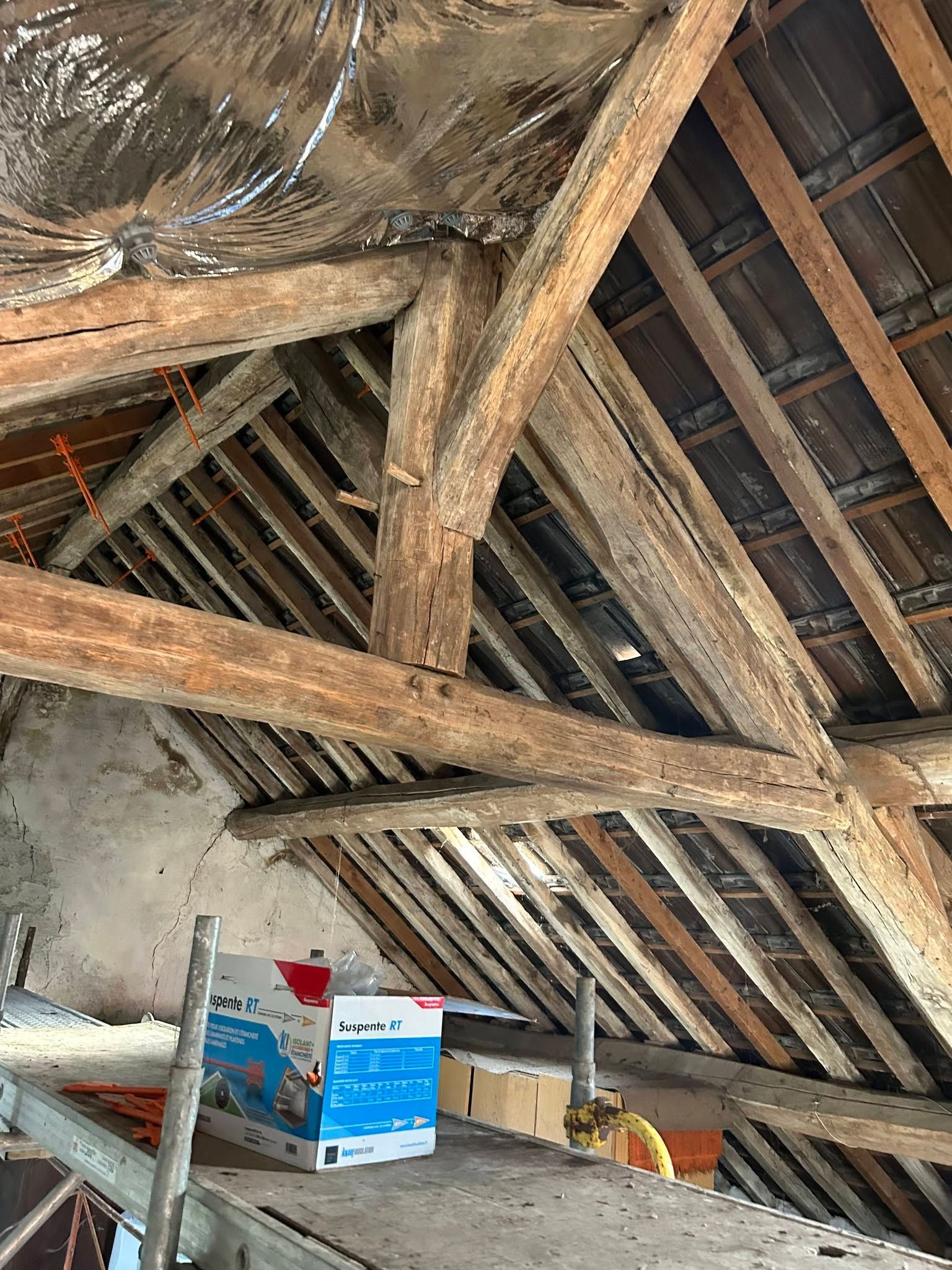 Chevrons en bois apparents dans les combles, avec l’isolation visible et des cartons entreposés sous un toit en pente.