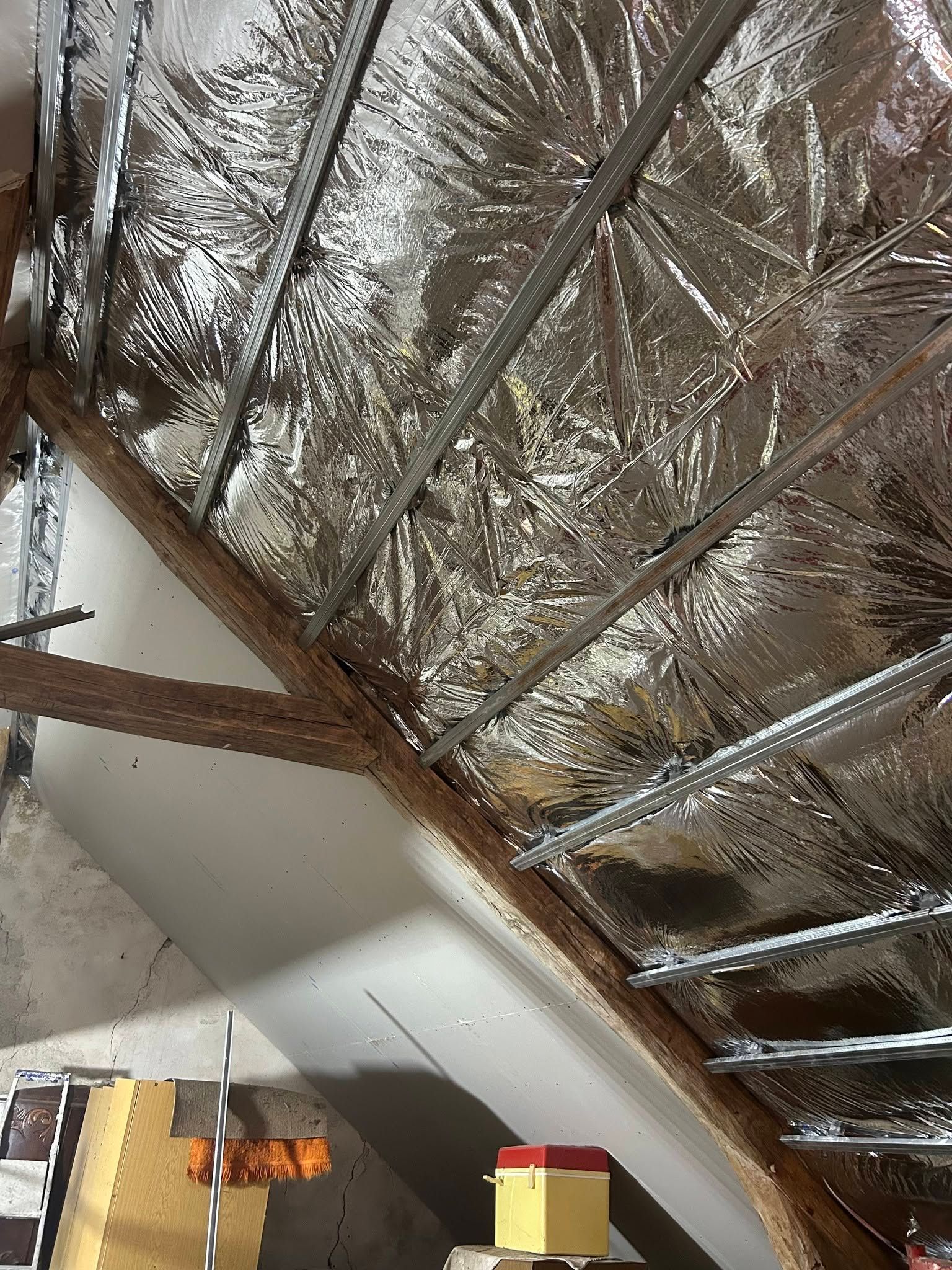 Plafond apparent avec isolant réfléchissant et poutres en bois au-dessus d’une cage d’escalier, avec quelques objets entreposés en dessous.