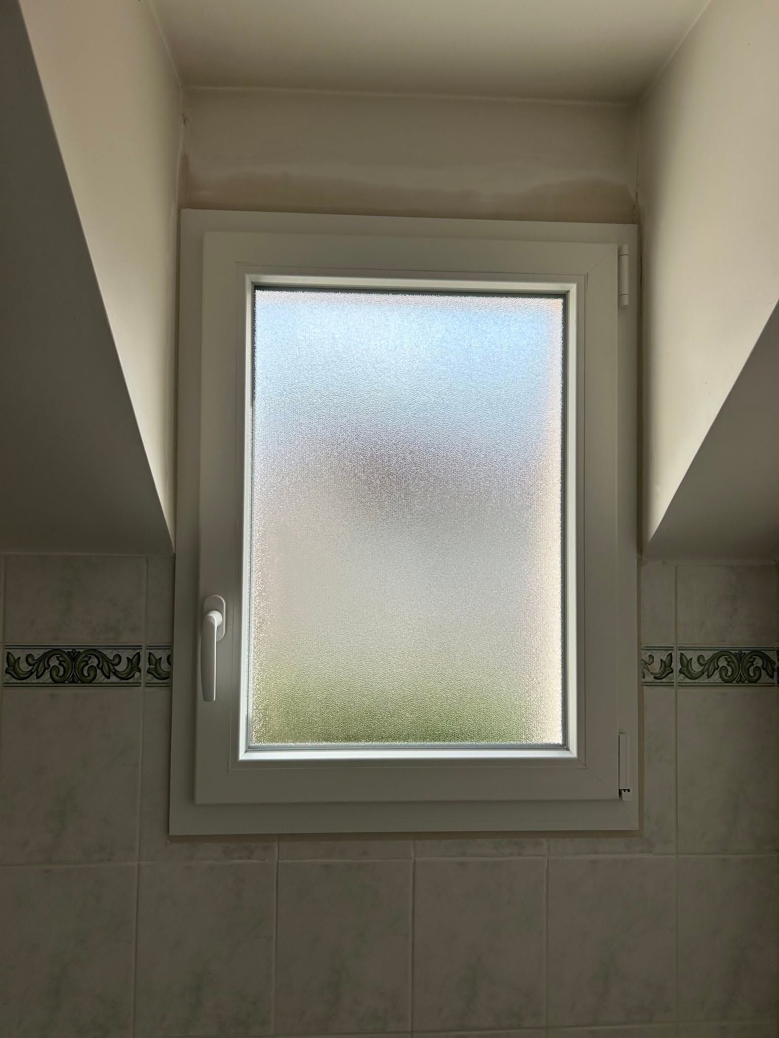 Fenêtre de salle de bains dépolie encadrée de blanc, entre des murs en pente et une crédence carrelée.