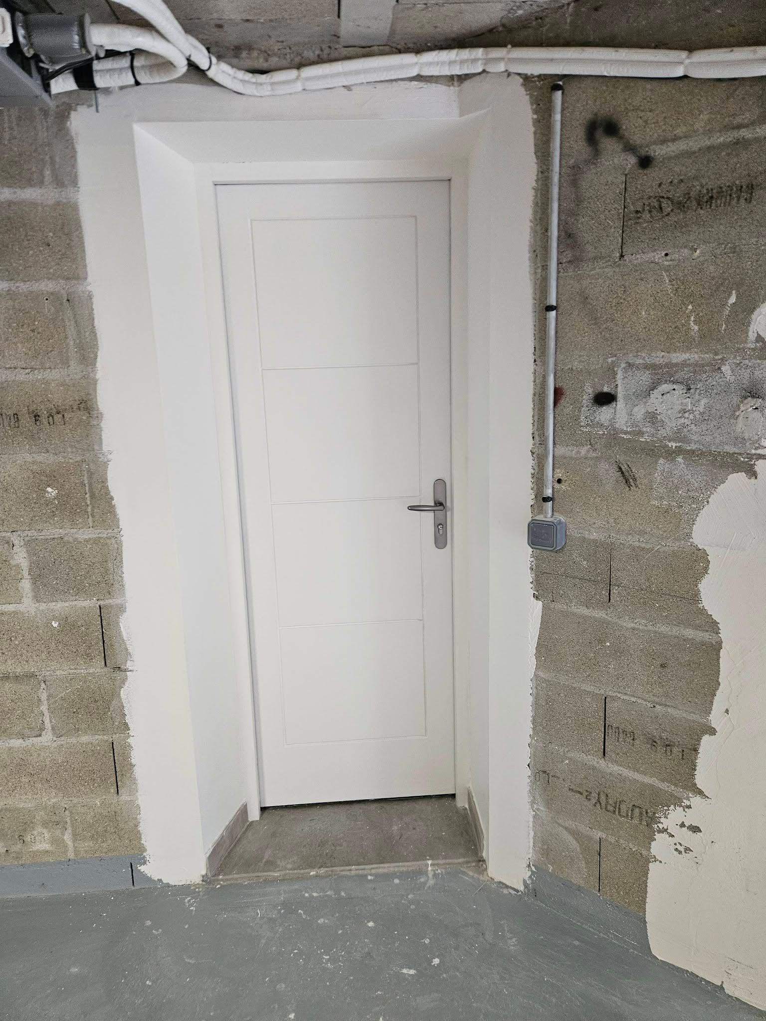 Porte blanche dans un mur de pierre brut non fini, avec du plâtre apparent et un sol en béton.