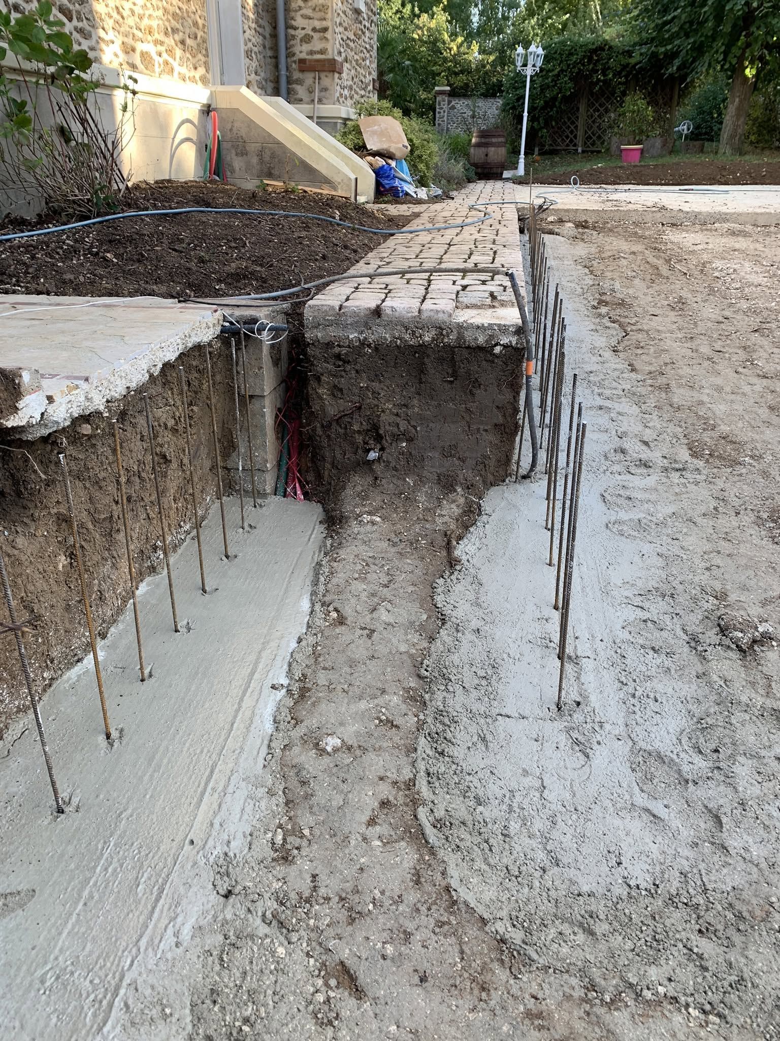 Tranchée de fondation en béton à nu sur un chantier, avec des armatures et un chemin de terre entre les murs.