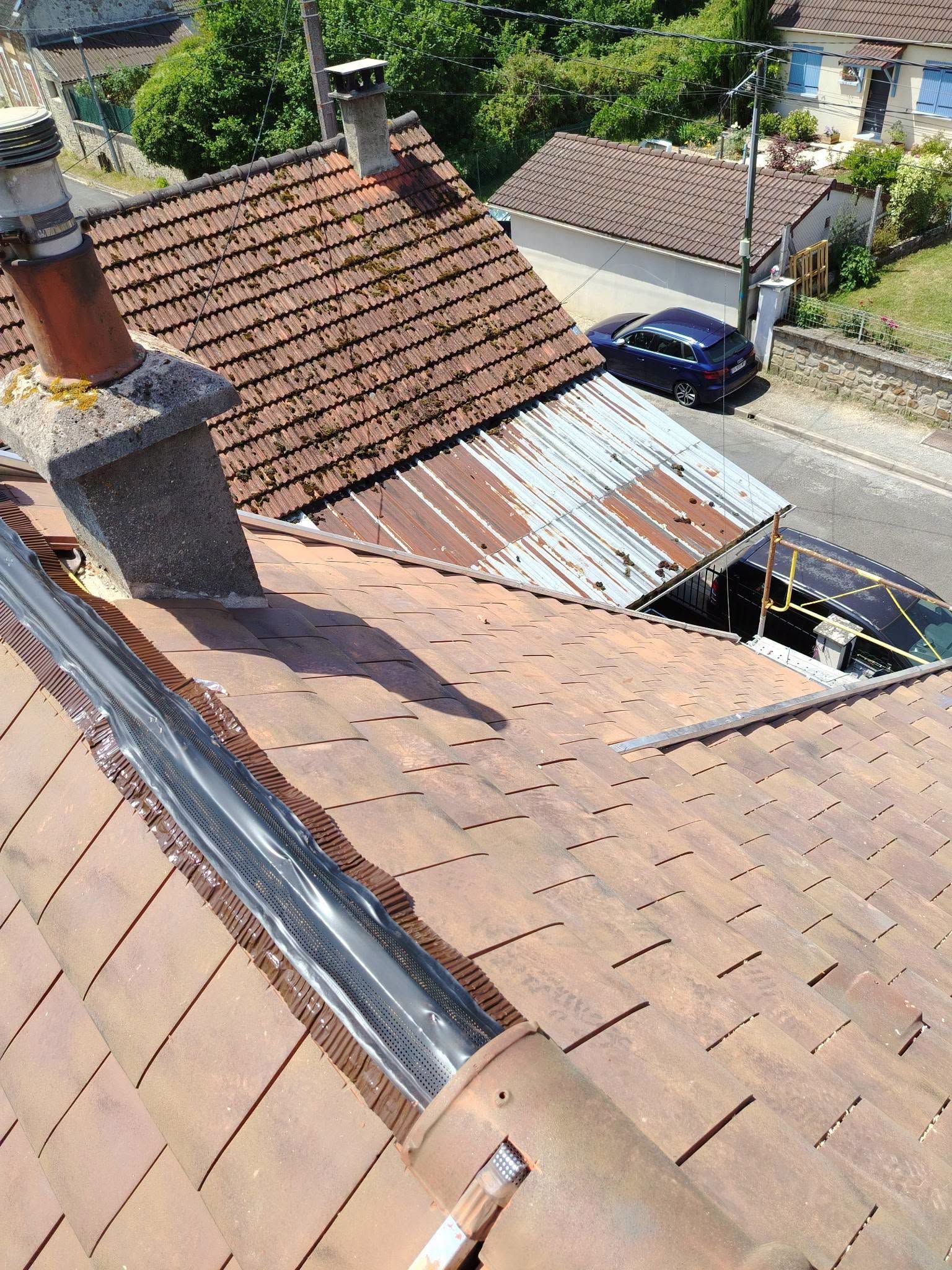Toiture avec des tuiles endommagées et une zone à découvert près d’une cheminée, surplombant une rue où se trouve une voiture bleue en contrebas.