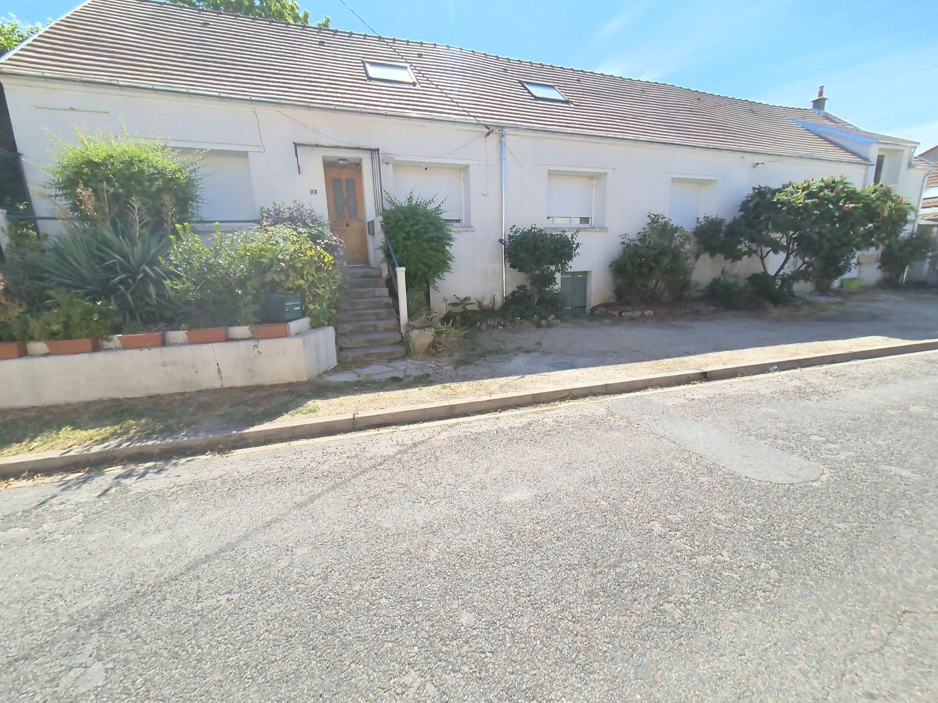 Maison blanche au toit de tuiles, avec un jardin devant, le long d’une route tranquille.