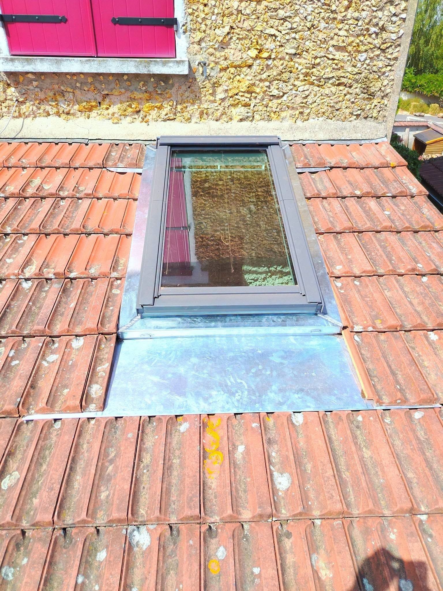 Fenêtre de toit avec une lucarne vitrée inclinée intégrée à une toiture en tuiles rouges, sous un mur recouvert de gravier.
