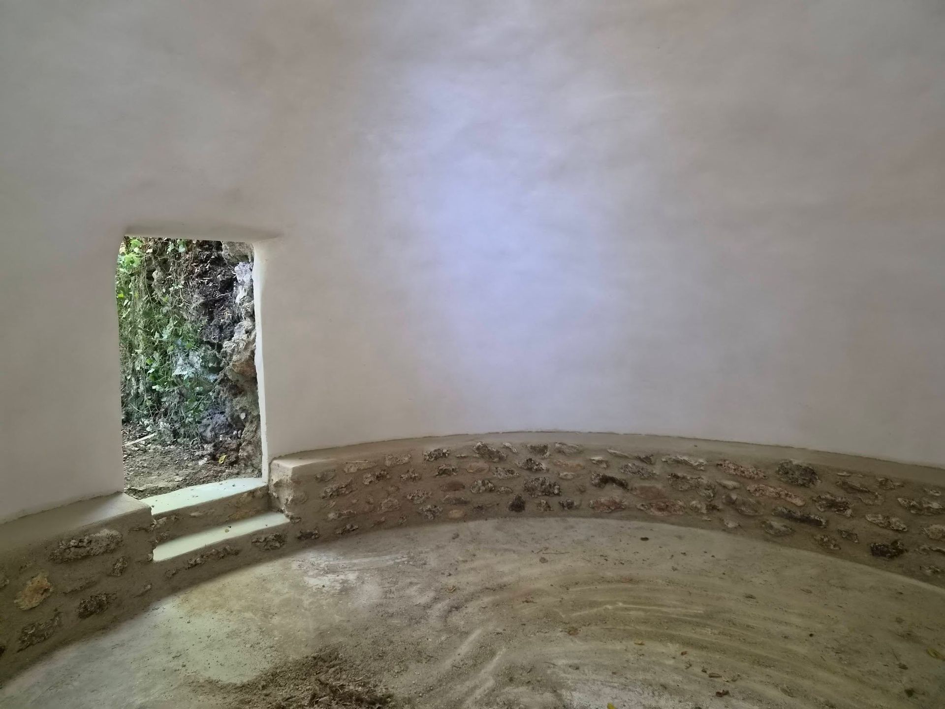 Pièce circulaire aux murs de pierre, avec une petite porte ouvrant sur la verdure et des marches à l’extérieur.