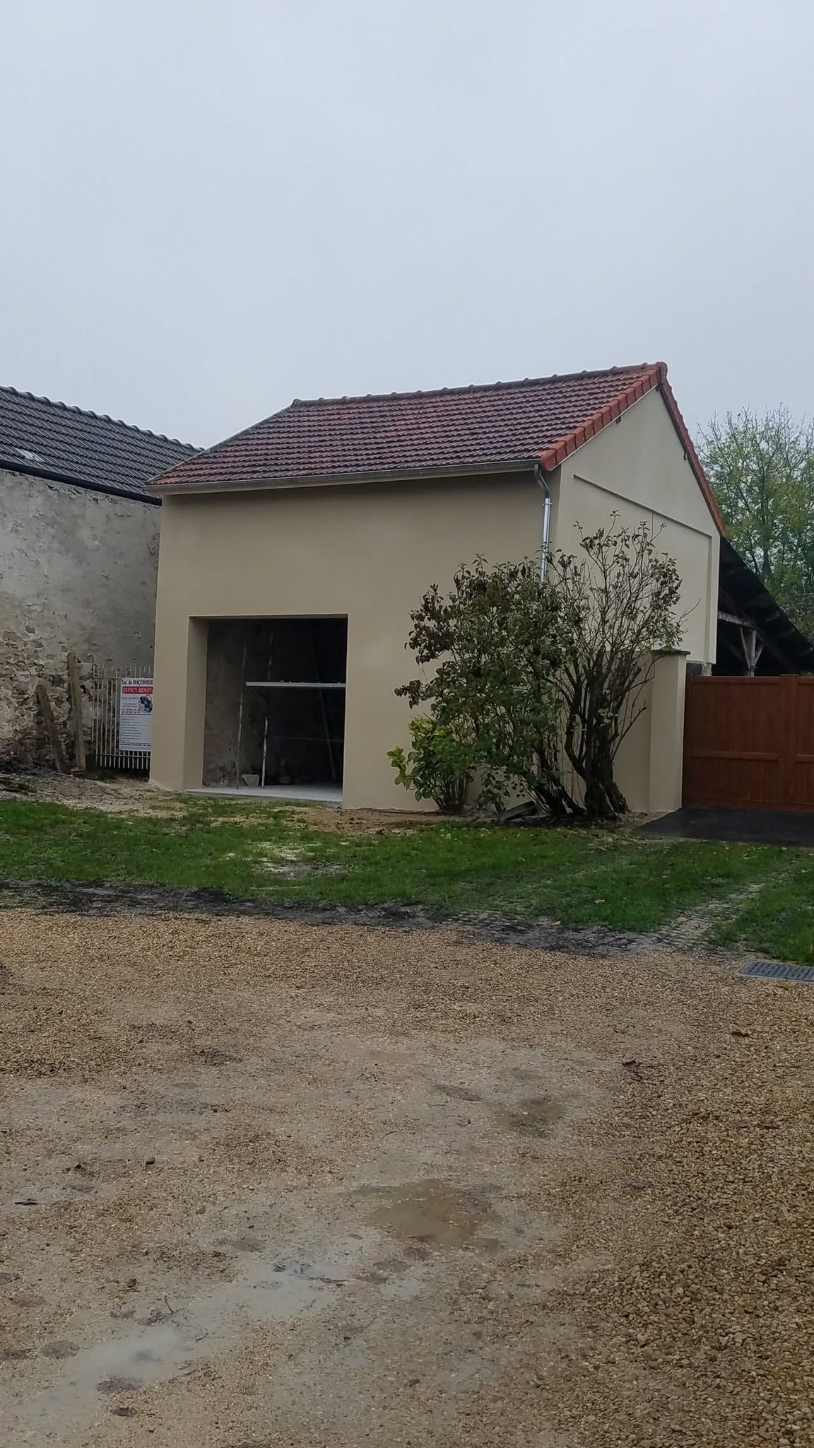 Petite maison inachevée au toit rouge, avec un garage ouvert dans une cour gravillonnée.