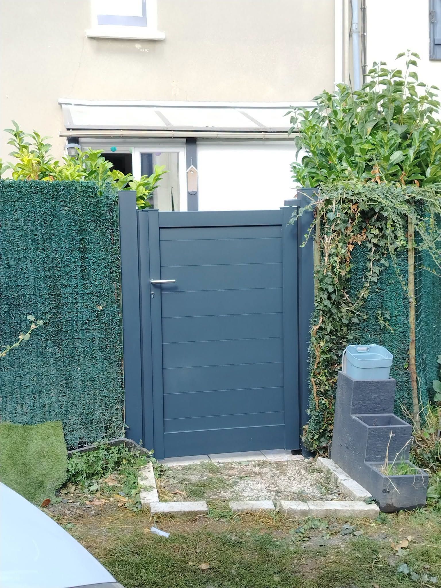 Portail de jardin bleu installé dans une haie, près du mur beige d’une maison et d’une fenêtre.