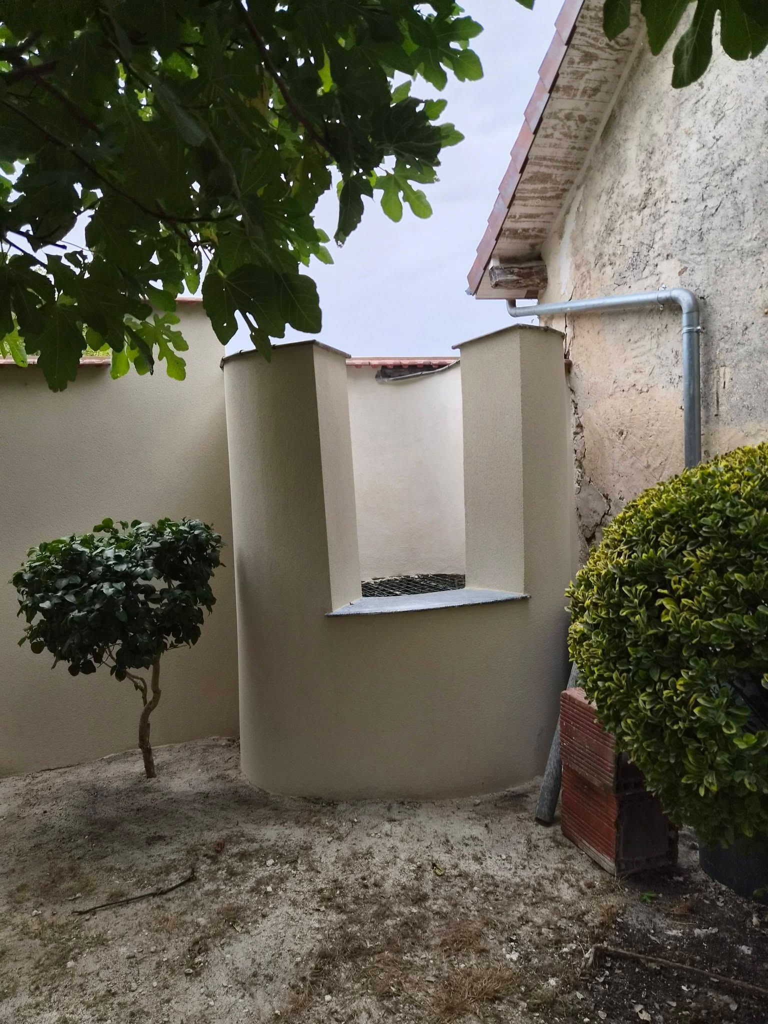 Petite cour avec des murs en stuc beige, une jardinière basse, un sol en gravier et des arbres et arbustes taillés.