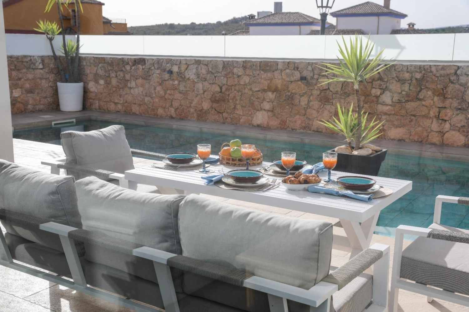 Comedor al aire libre junto a una piscina, con una mesa puesta, asientos grises y plantas en macetas al atardecer