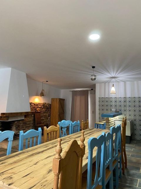 Comedor con una larga mesa de madera, sillas azules y amarillas y una chimenea de piedra bajo lámparas colgantes.