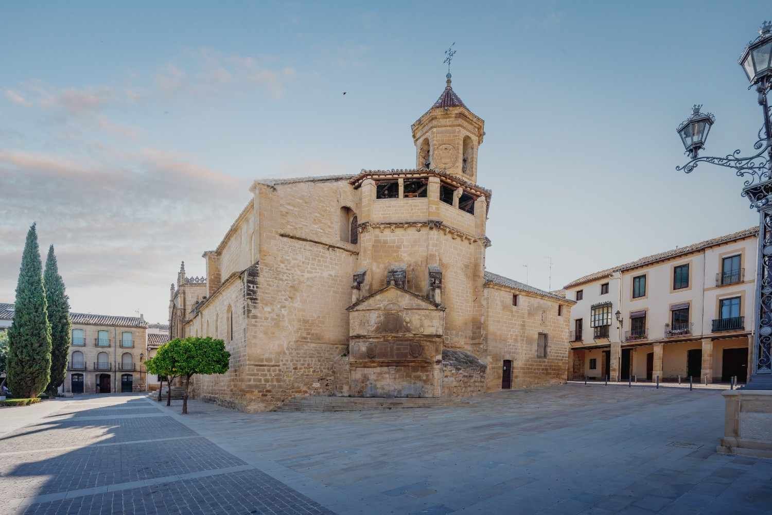 Iglesia histórica de piedra en una plaza tranquila, con edificios antiguos y el cielo del atardecer al fondo