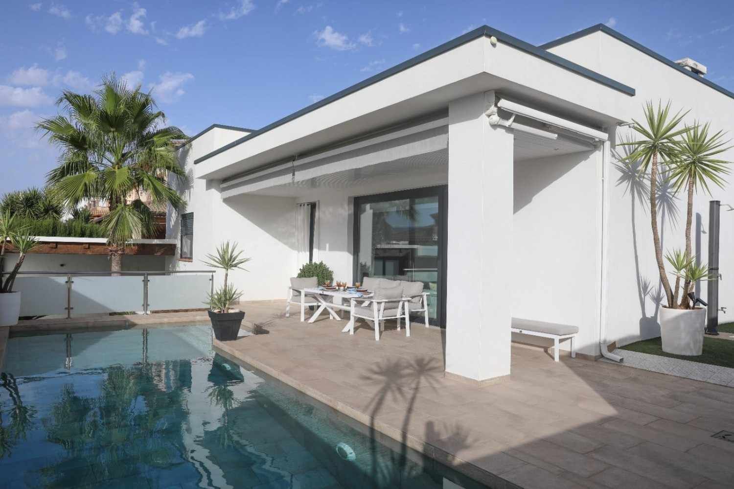 Patio moderno de una casa blanca con piscina, mesa de comedor al aire libre y palmeras en macetas