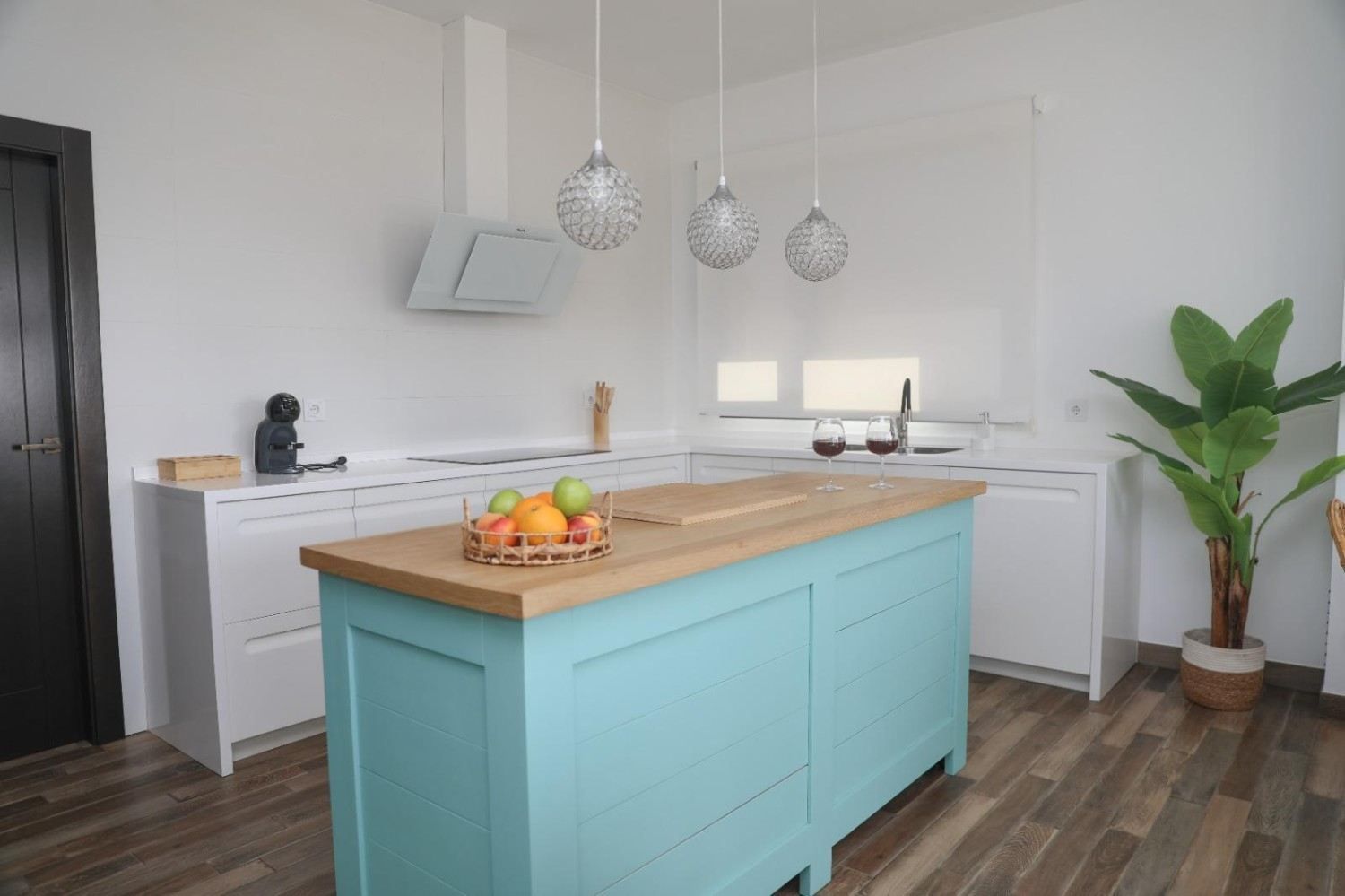 Cocina luminosa con una isla turquesa, armarios blancos y una planta en maceta junto a la ventana.