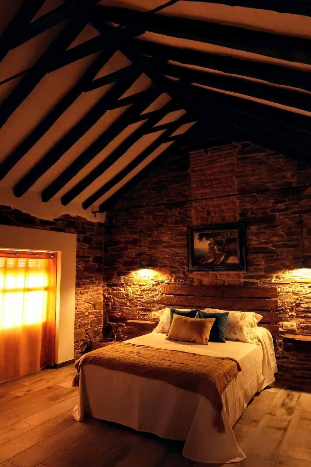 Dormitorio rústico con una cama, paredes de ladrillo visto, iluminación cálida y techo con vigas de madera