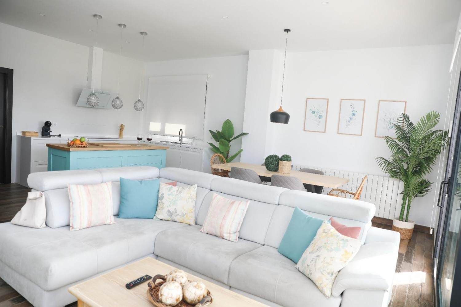 Luminoso salón con un sofá seccional blanco, cojines en tonos pastel, una mesa de madera y una cocina abierta.