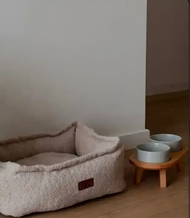 Cama beige para mascotas junto a un pequeño comedero de madera con dos cuencos plateados sobre un suelo de madera.